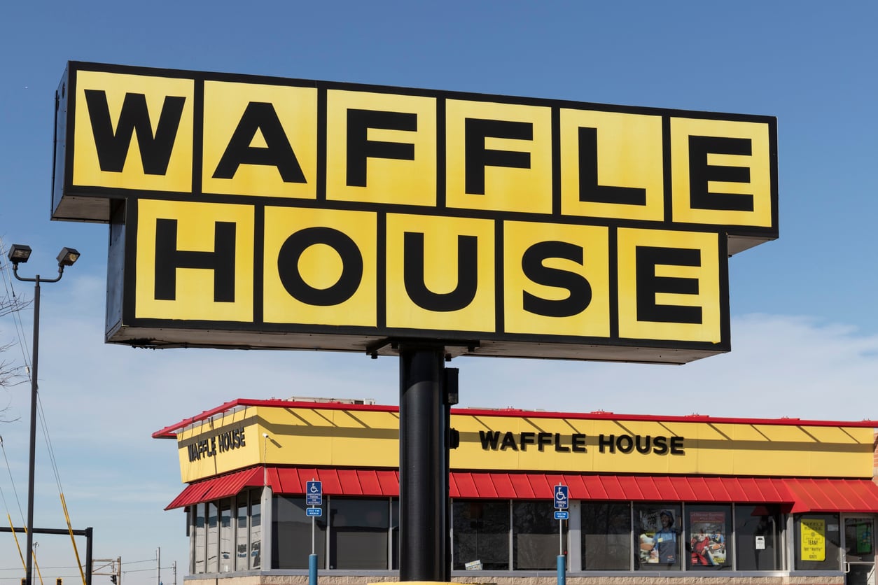 Large yellow Waffle House sign with black letters in front of a Waffle House restaurant building, featuring a red roof and blue sky in the background.