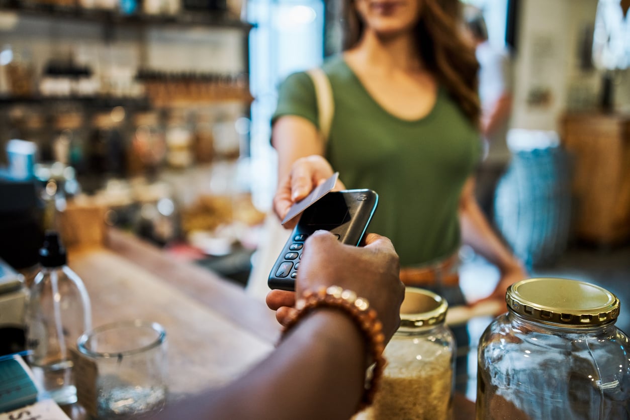 A woman in a green shirt holds out a card to pay at a store counter, where another person holds a card reader. Glass jars are visible on the counter.