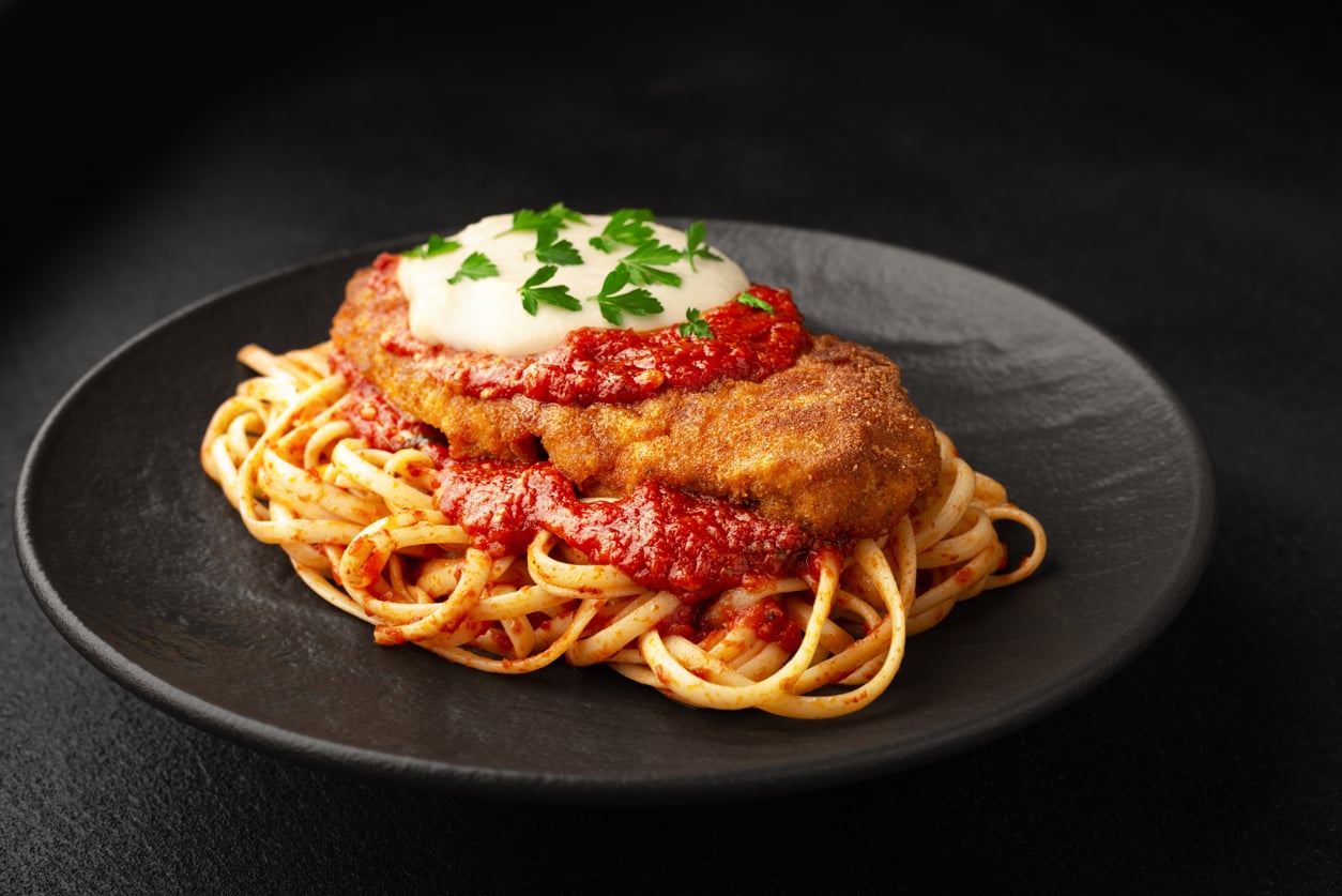 A black plate with spaghetti topped with marinara sauce, breaded chicken cutlet, melted cheese, and chopped parsley, set on a dark background—one of those comforting meals middle-class moms made for family dinners.