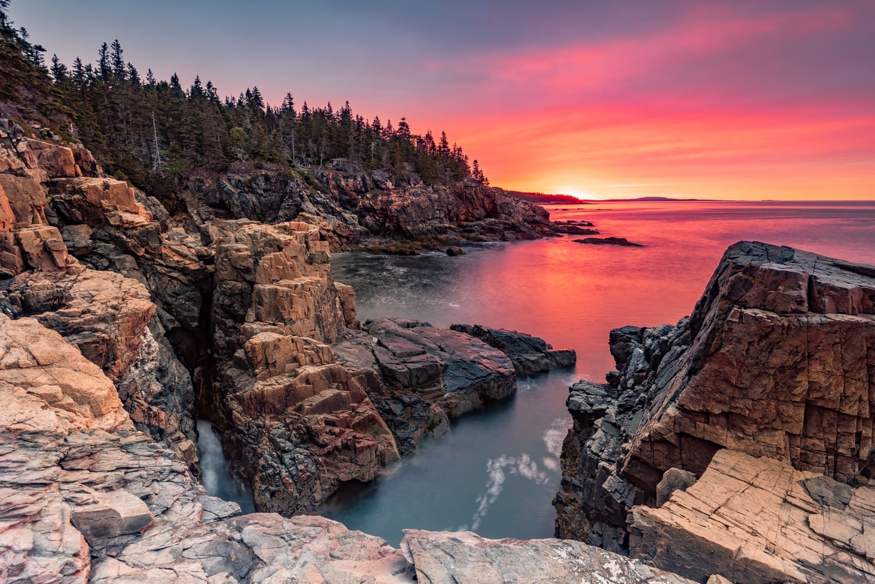 Rocky coastline with pine trees at sunset, vibrant pink and orange sky reflected on calm water, dramatic cliffs and distant horizon create a serene, scenic view—an idyllic choice among top vacation spots.