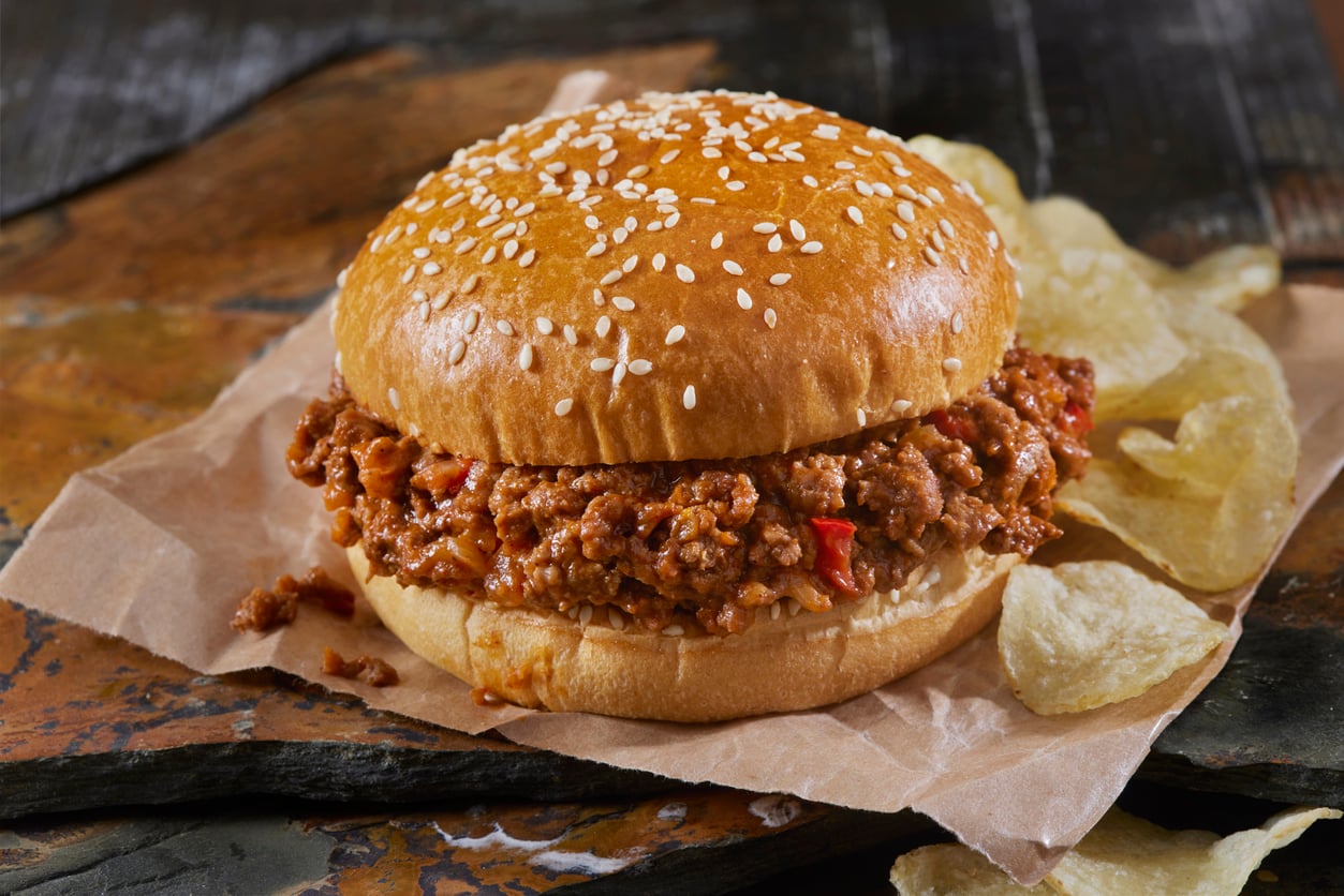 A sloppy joe sandwich with a sesame seed bun sits on brown parchment paper next to potato chips. The filling is a saucy ground meat mixture with visible bits of red pepper.