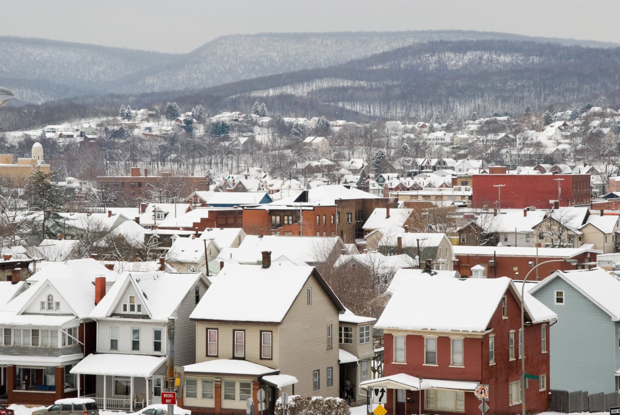 Snow-covered houses and buildings in a small town with mountains in the background. Leafless trees are scattered throughout, and the scene appears quiet on a winter day.