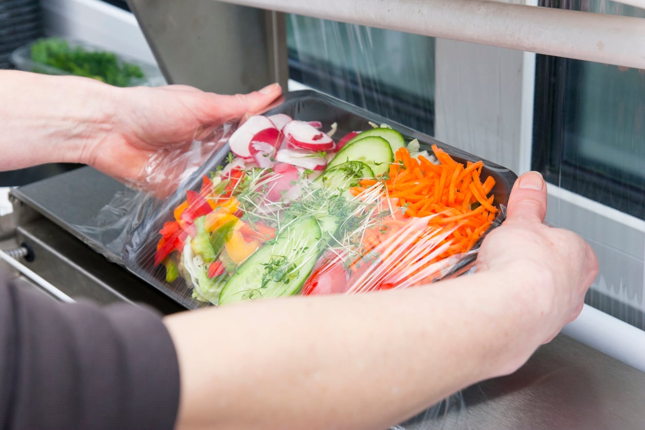 Hands wrapping a tray of assorted fresh vegetables, including sliced radishes, cucumbers, carrots, and bell peppers, with clear plastic wrap in a kitchen setting.