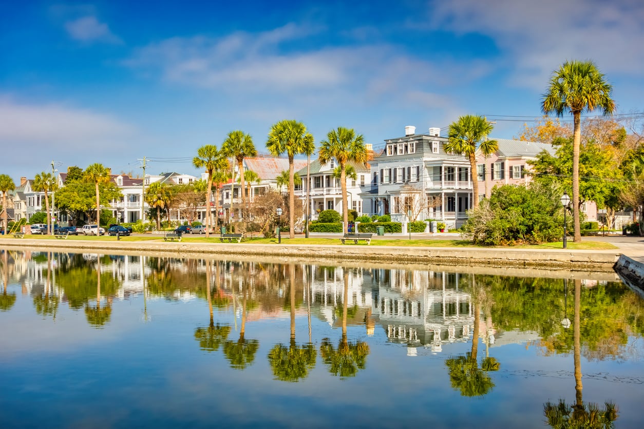 Colorful historic houses and palm trees line a waterfront street, reflected clearly in the calm water under a bright blue sky with some scattered clouds—a serene scene that invites peaceful strolls, especially cherished after age 50.