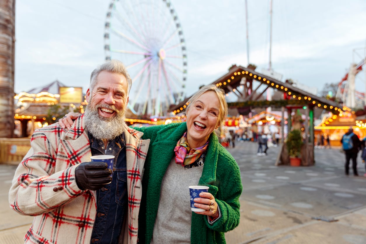 Smiling older couple enjoying hot drinks at an outdoor fair, with bright lights and a Ferris wheel in the background. The atmosphere is lively and festive.
