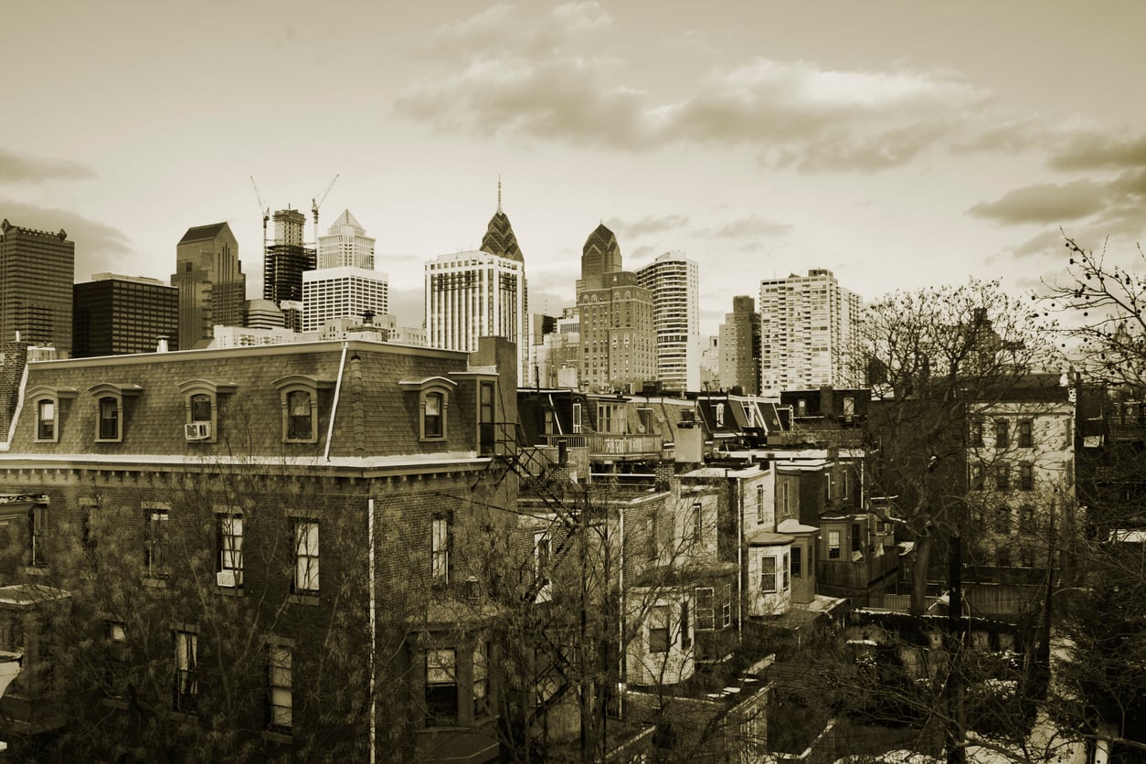 Sepia-toned photo of a city skyline with modern skyscrapers behind older brick rowhouses and leafless trees in the foreground.