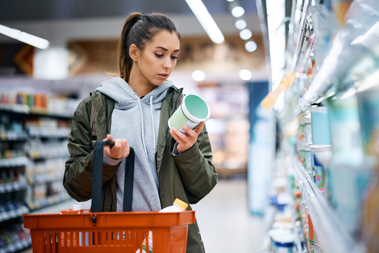 A woman with a shopping basket examines a product label while standing in a grocery store aisle, surrounded by shelves stocked with various items.