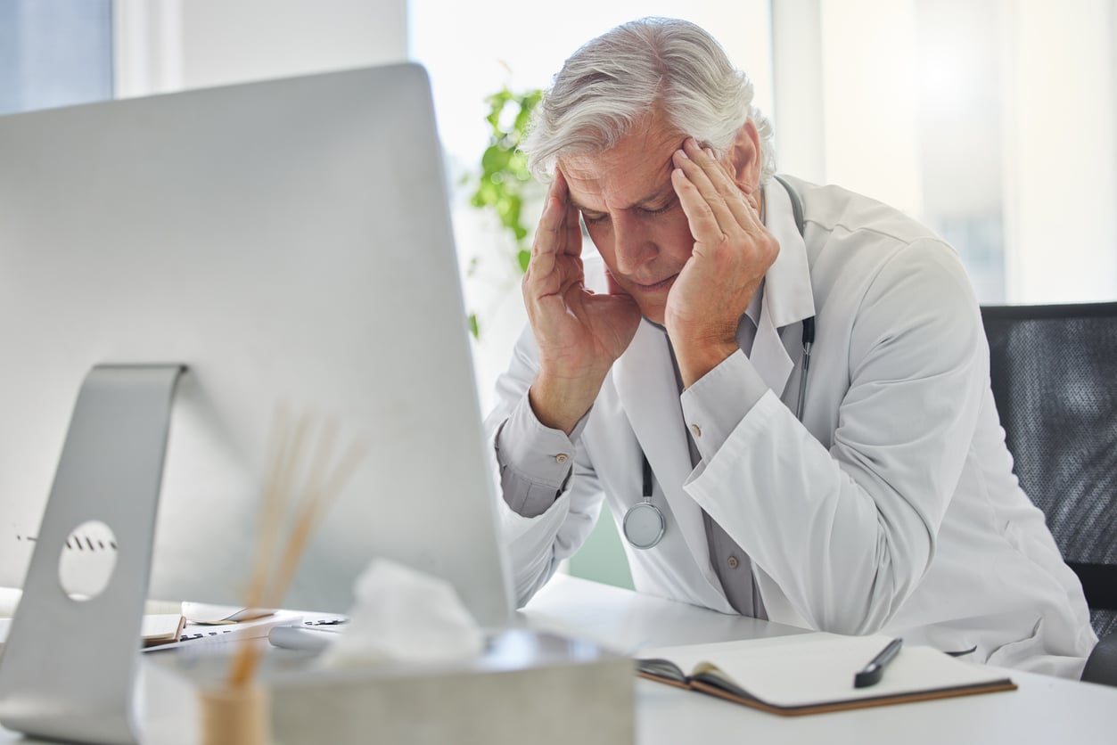 A doctor in a white coat sits at a desk, looking stressed with hands on his temples, in front of a computer and an open notebook—perhaps contemplating retirement tips as he manages the daily pressures of his demanding profession.