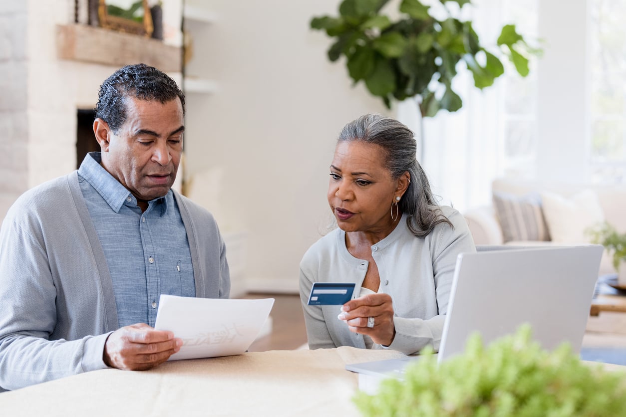 An older couple sits at a table, looking at documents and a credit card, with a laptop open in front of them. They appear to be focused and discussing something important.