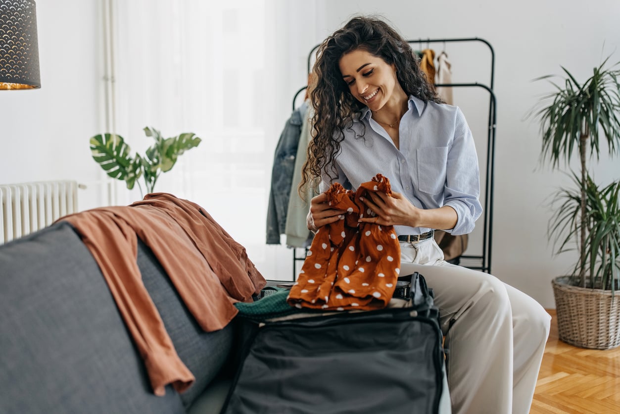 A woman smiles while packing clothes into a suitcase in a bright, modern living room. There are garments on the sofa and a clothing rack with hanging items in the background.