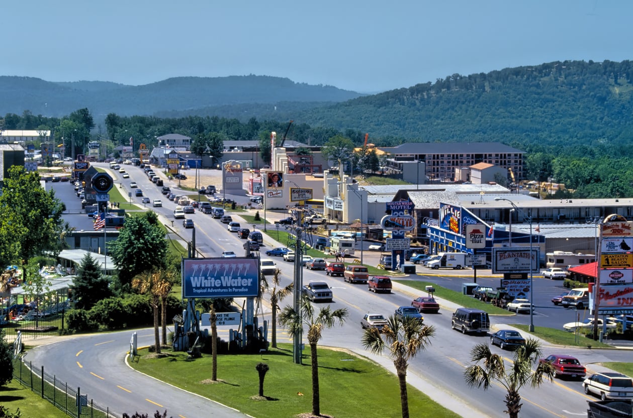 A busy street in a tourist town with many cars, palm trees, billboards, shops, and attractions—now counted among the travel spots Boomers are learning to love—surrounded by green hills under a clear blue sky.