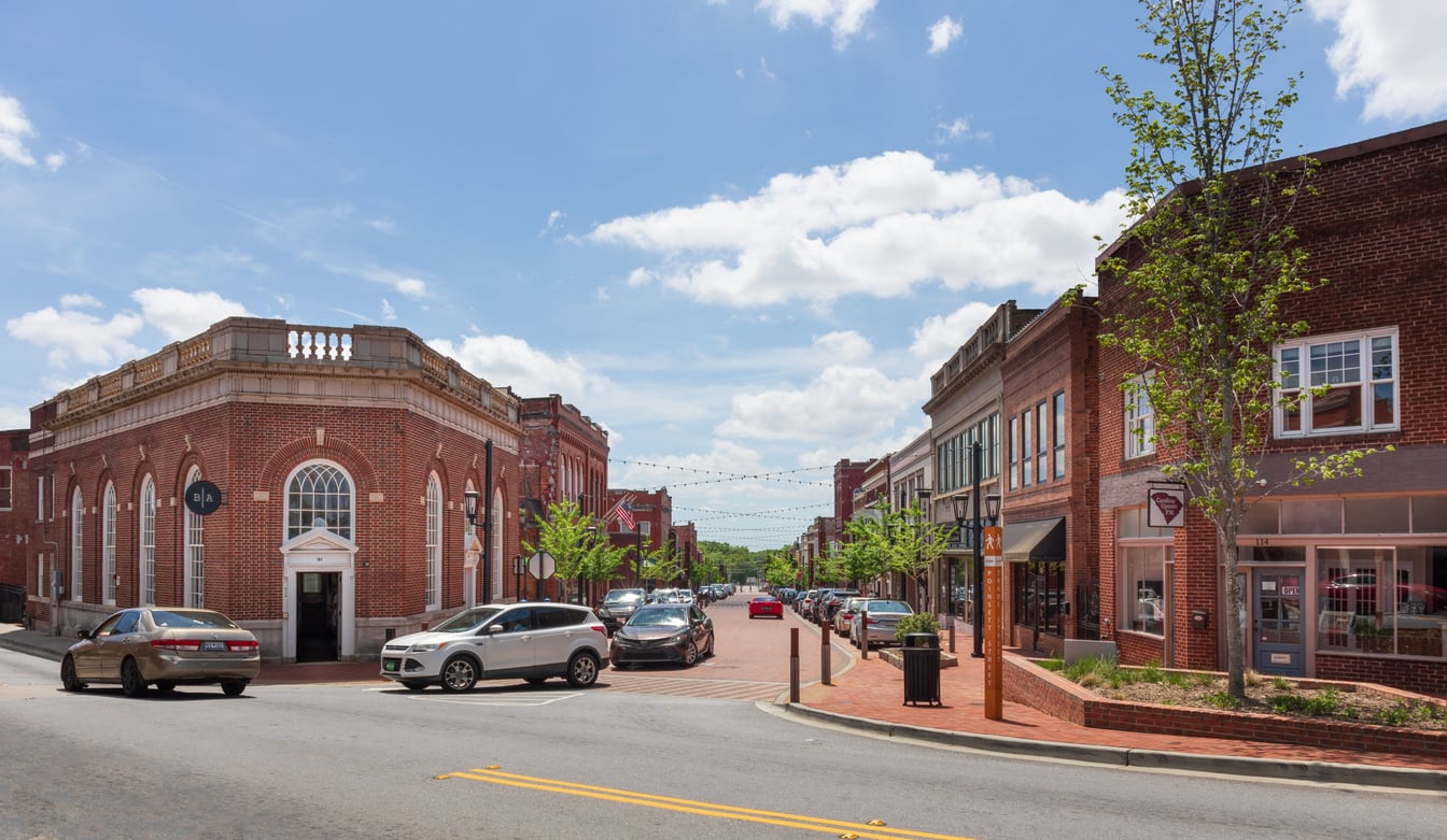 Sunny day on a small town street with red brick buildings, cars parked along the road, string lights overhead, and a few young trees lining the sidewalks.