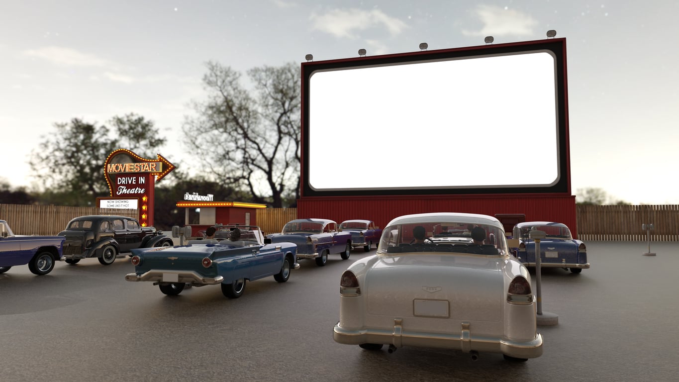 Several classic cars are parked at a drive-in theater in front of a large blank movie screen at dusk, with a neon sign and trees in the background.