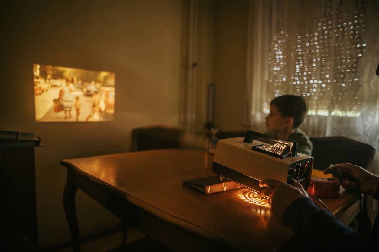 A vintage slide projector displays a family photo on the wall. A child sits at a table, watching, while an adult operates the projector in a warmly lit room with lace curtains.