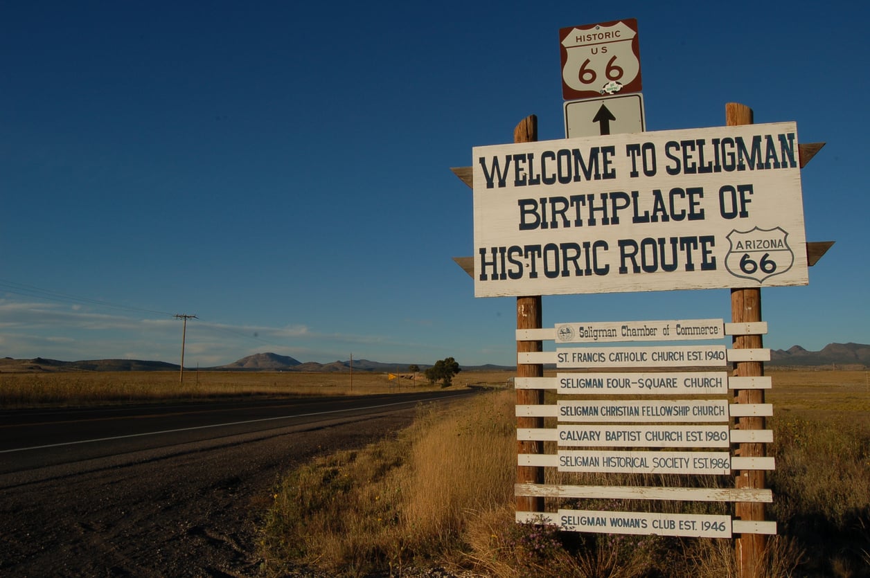 A wooden sign by a rural road welcomes visitors to Seligman, birthplace of Historic Route 66 and one of America’s most nostalgic travel destinations, listing local organizations. Fields and distant mountains are visible under a clear sky.
