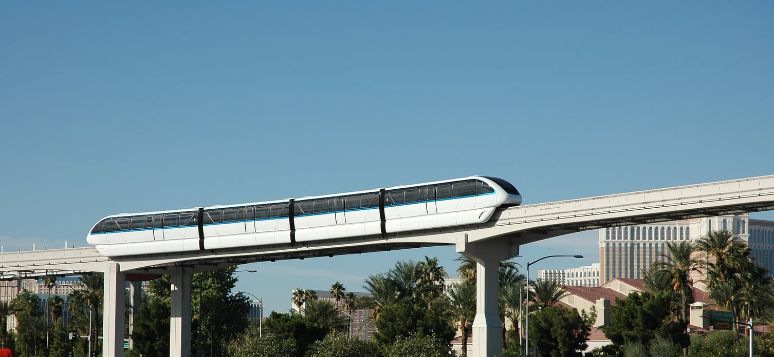 A white monorail train with blue stripes travels on an elevated track above palm trees and buildings under a clear blue sky, highlighting one of the unique sights you’ll encounter while discovering fascinating facts about Las Vegas.