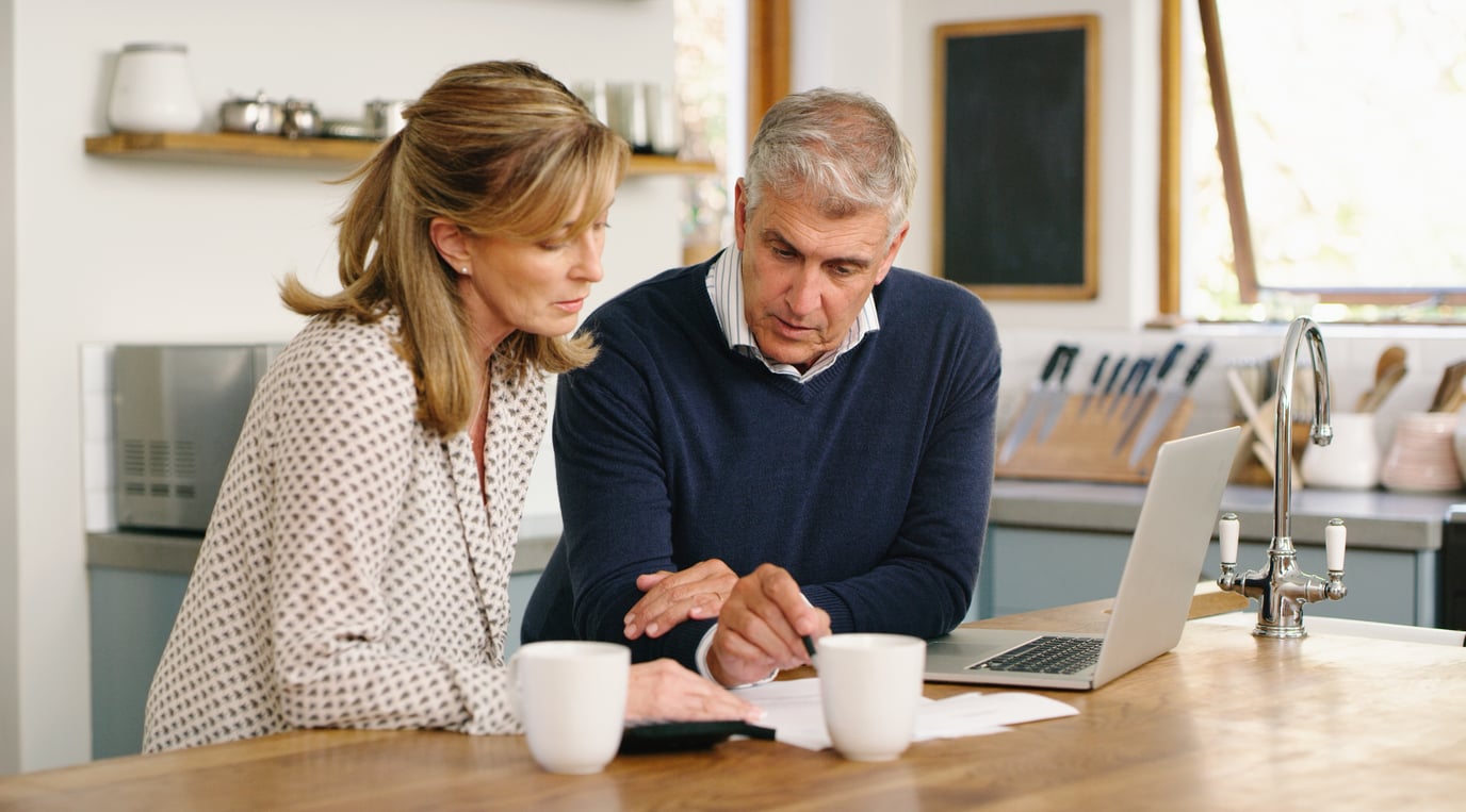 A middle-aged couple sits at a kitchen counter, looking at documents together. A laptop and two mugs are on the counter. As the man points at the papers and the woman listens, they discuss their travel behaviors for an upcoming trip.