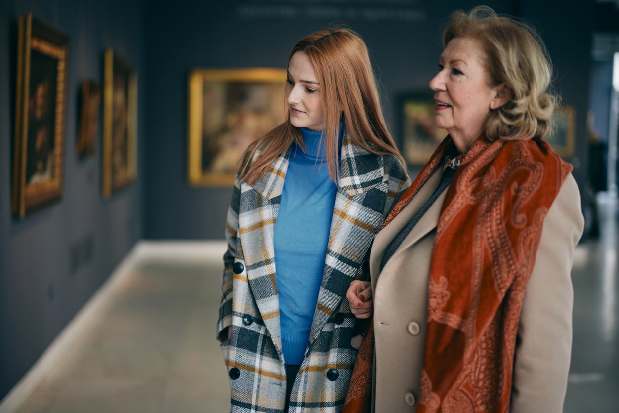 Two women in coats stand side by side, looking at artwork on the walls of an art gallery. The setting is bright and modern, with several framed paintings displayed in the background.