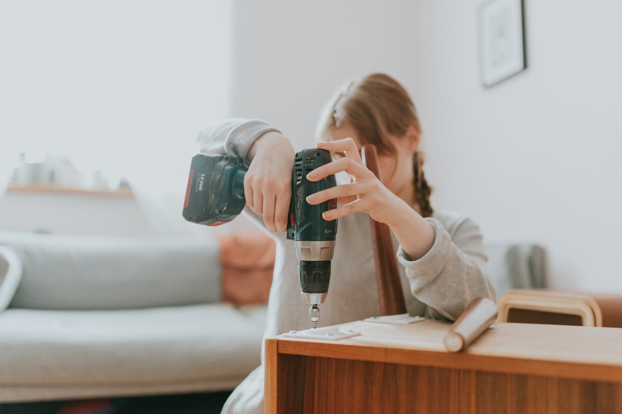 A person uses a cordless power drill to assemble wooden furniture in a cozy room with a sofa in the background. Their face is partially hidden by their arm and hair.