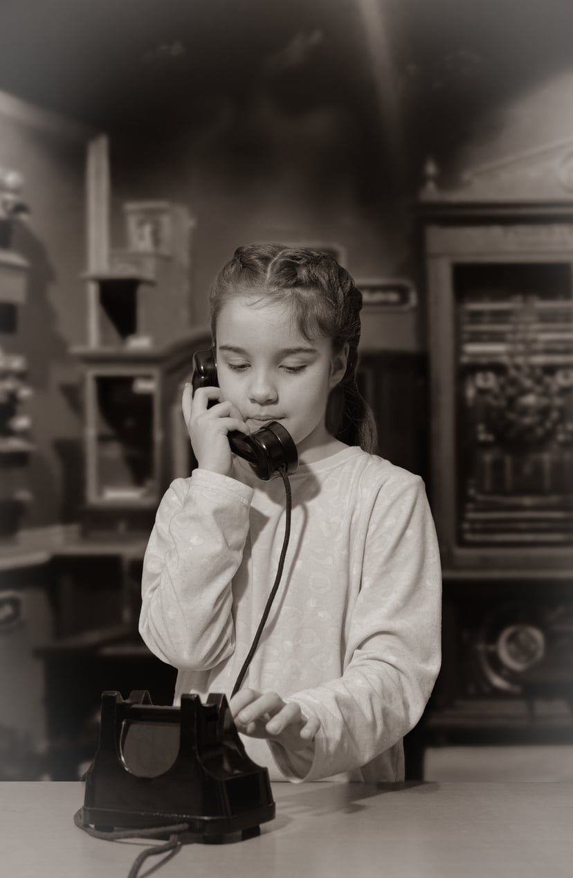 A young girl with braided hair uses a vintage rotary phone, holding the receiver to her ear and dialing with her other hand. The background appears to be an old-fashioned room. The image is in sepia tones.