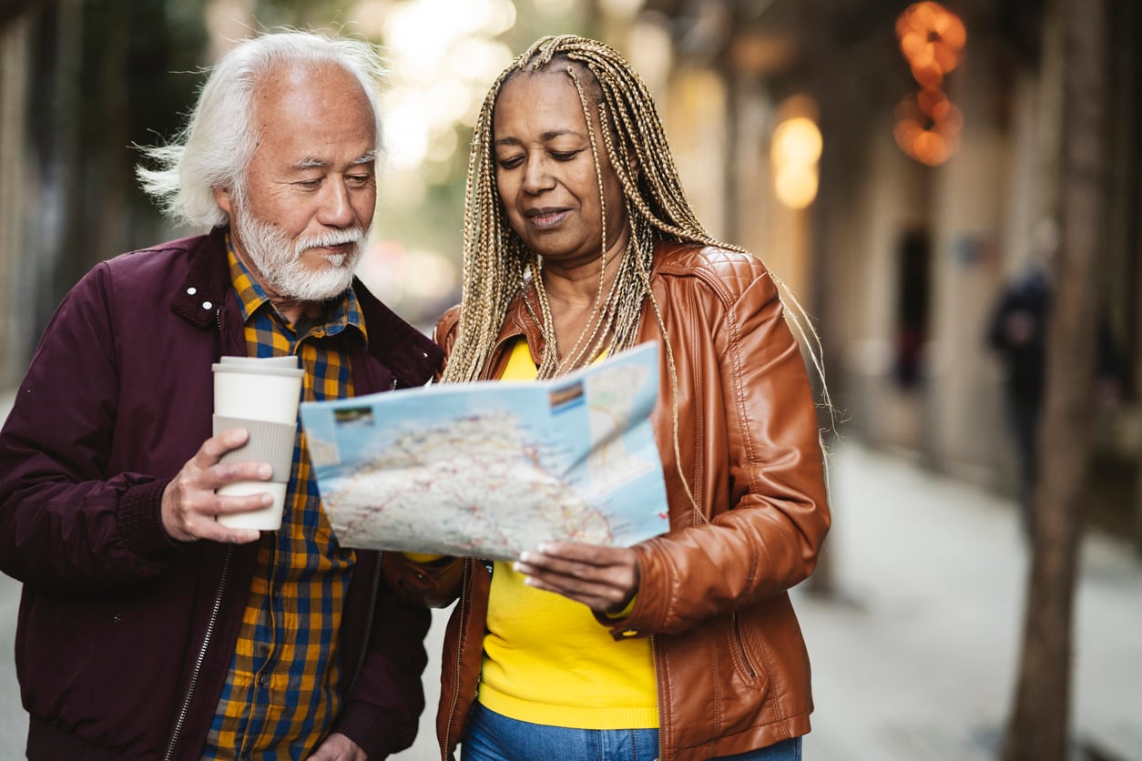 An older man holds a coffee cup while standing next to a woman reading a map. They appear to be exploring a city street together, showcasing their travel habits as they navigate new surroundings side by side.