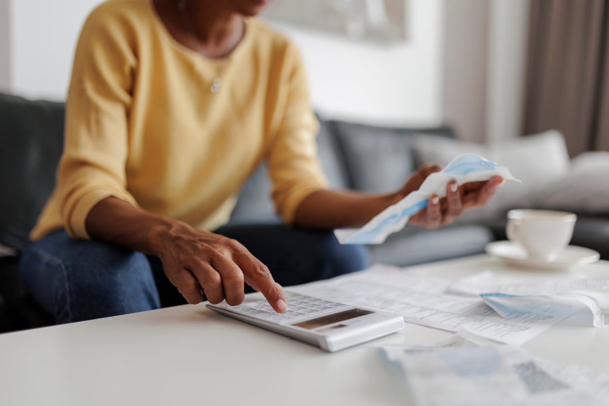 A person in a yellow sweater sits on a couch, using a calculator and holding receipts, with papers and a coffee cup on a white table, appearing to manage finances.