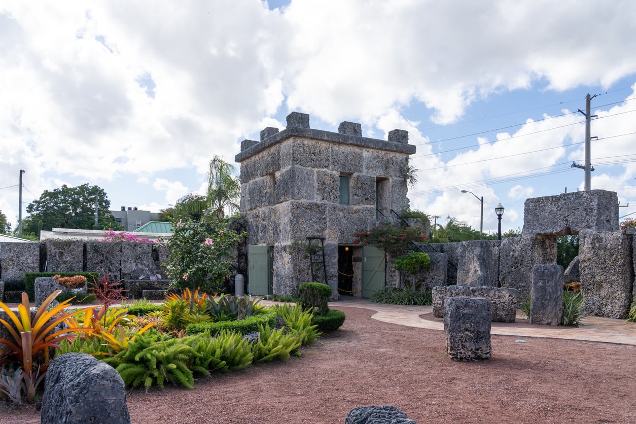 A stone castle-like structure with battlements stands in a garden with tropical plants, flowering bushes, and pathways under a partly cloudy sky, capturing the charm of Classic Roadside Attractions.