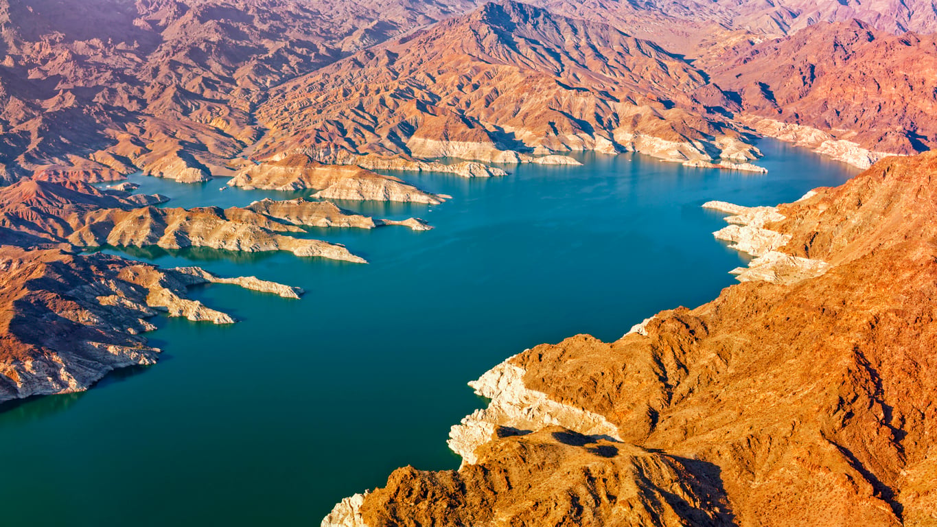 Aerial view of a winding blue lake surrounded by rugged, sunlit brown mountains and rocky shoreline, with visible water level marks on the slopes—an iconic landscape near Las Vegas that highlights some interesting facts about Las Vegas and its surrounding natural beauty.