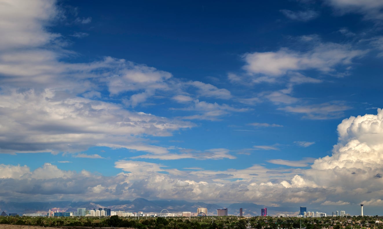 A wide view of a city skyline under a blue sky with large, scattered clouds and distant mountains in the background—an iconic scene that brings to mind fascinating facts about Las Vegas and its vibrant metropolitan landscape.