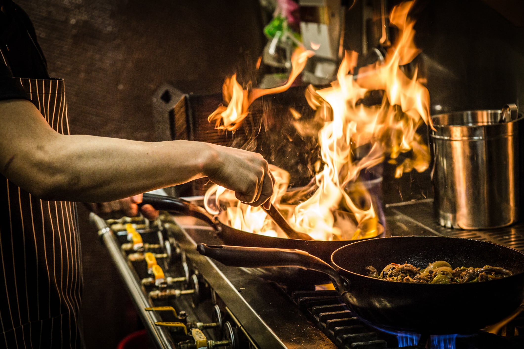 A person stirs food in a frying pan over a stove with tall flames rising, creating a dramatic cooking scene in a kitchen.