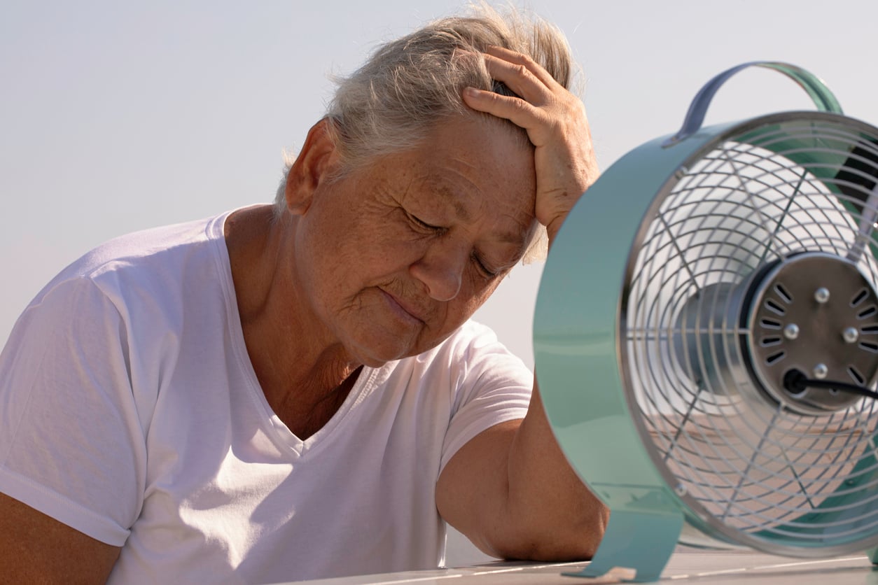 An older person in a white shirt sits with eyes closed, looking tired, resting their head on their hand next to a small electric fan—perhaps reflecting on recent travel behaviors. The background is bright and sunny.