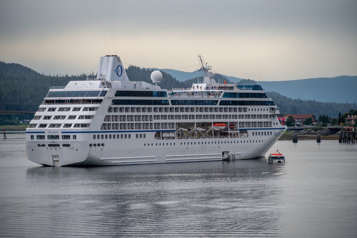 A large white cruise ship is docked near a forested shoreline, with a small tender boat nearby on the water under a cloudy sky.