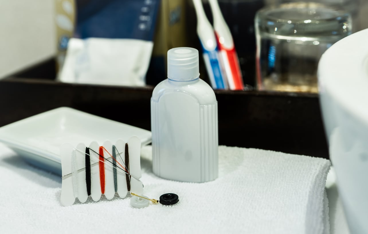 A small white toiletry bottle, sewing kit, and soap dish—classic things hotels used to offer guests—rest on a white towel in a bathroom, with toothbrushes and a glass visible in the background.