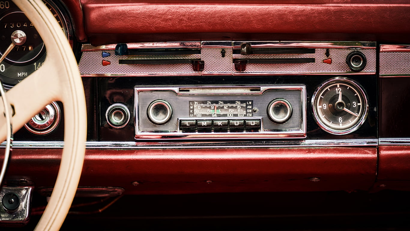 Close-up of a vintage car dashboard featuring an analog radio, a round clock on the right, speedometer on the left, and red upholstery.