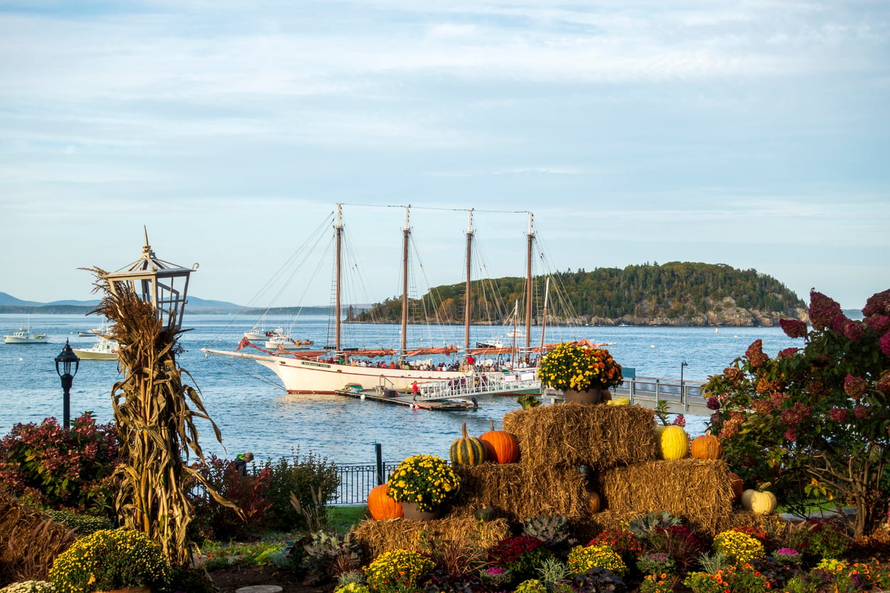 A sailboat on calm water near an island, with a foreground of autumn decorations—including hay bales, pumpkins, and flowers—in a garden by the shore, evokes the charming atmosphere often found in the best fall towns in America.