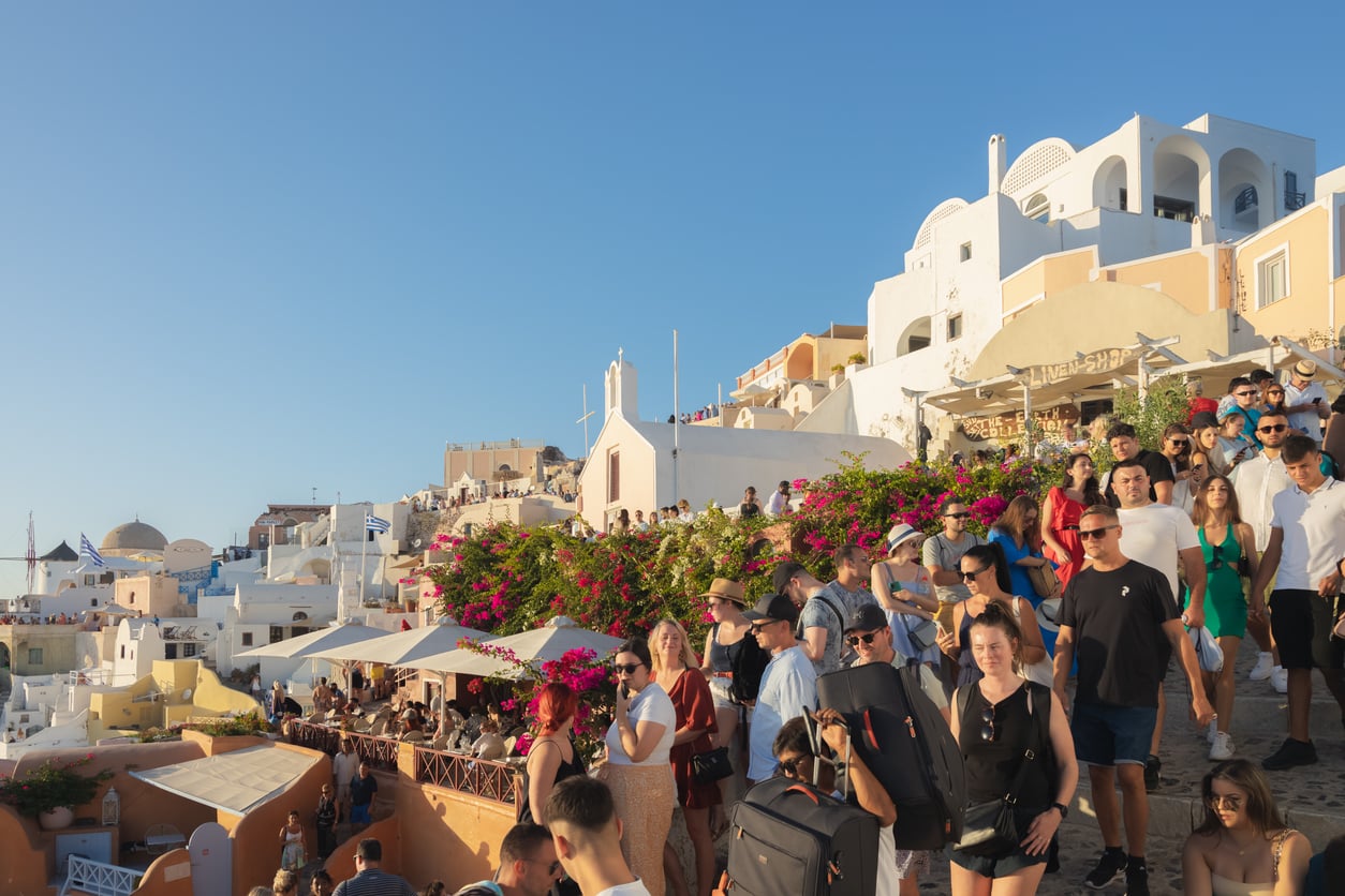 A large crowd of tourists gathers on a sunny day in front of colorful buildings and white houses on a hillside in Santorini, Greece. Bougainvillea flowers and clear blue sky are visible.