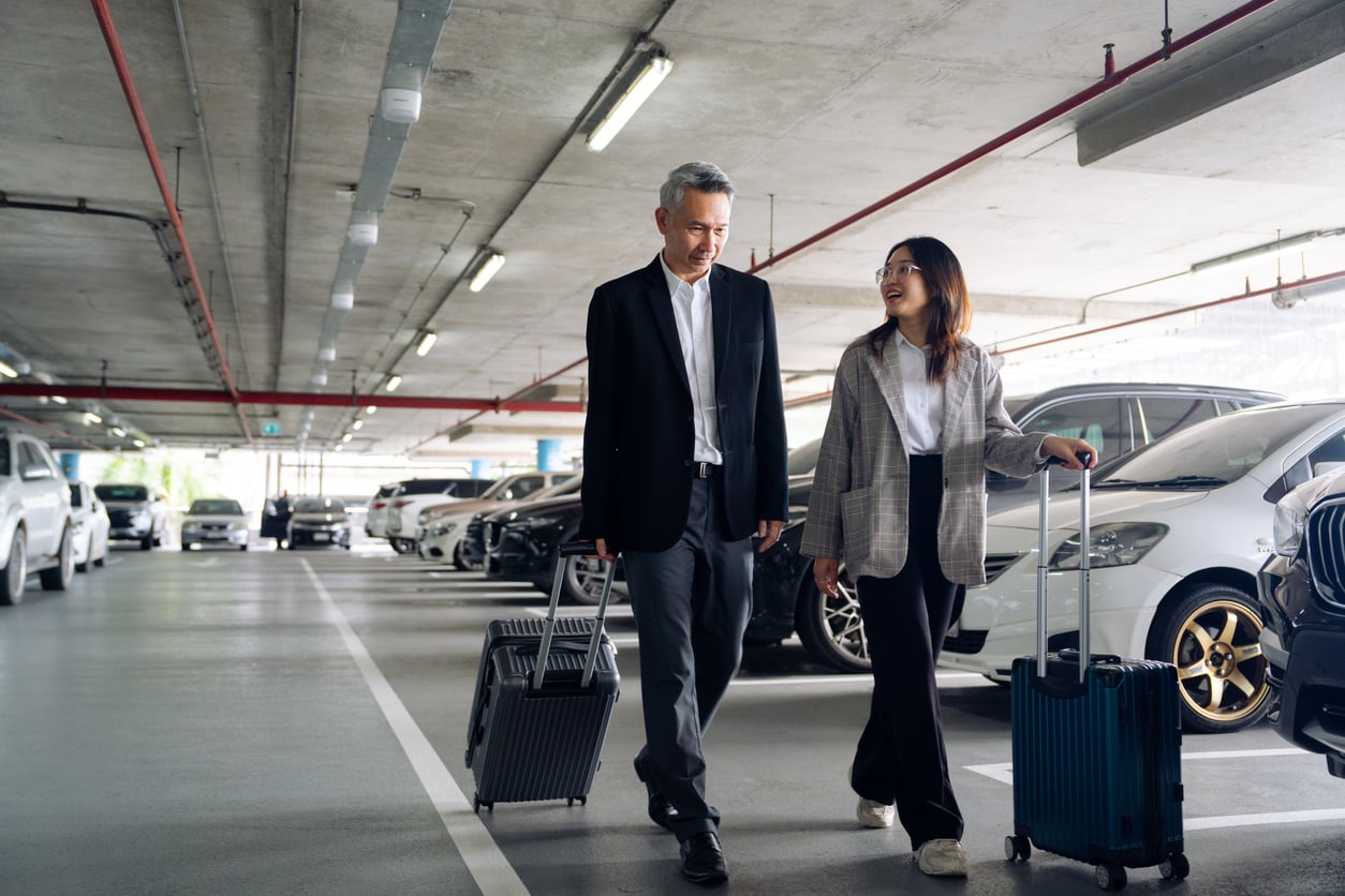 Two people walk together in a parking garage, each pulling a suitcase. They are both dressed in business attire and appear to be talking and smiling, surrounded by parked cars in the background.