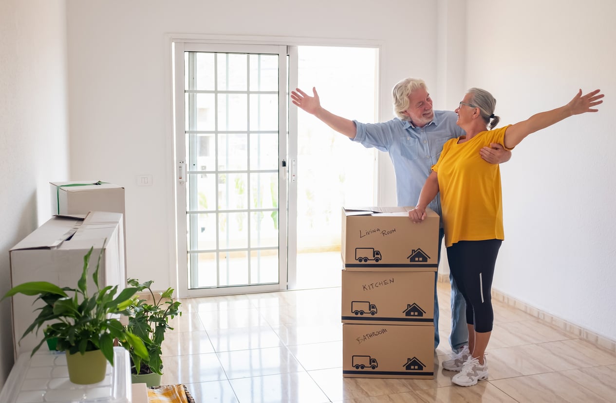 Two older adults happily embrace in a bright, empty room with moving boxes labeled “Living Room,” “Kitchen,” and “Bathroom.” Reflecting the spirit of Baby Boomer changes, they stand near large glass doors, smiling with arms outstretched as they welcome a new chapter together.