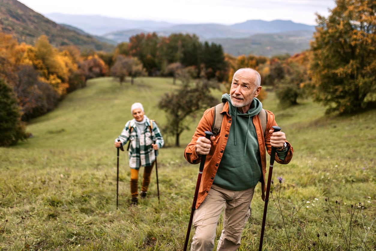 An older man and woman, demonstrating active travel behaviors, hike up a grassy hill using trekking poles, surrounded by autumn trees and distant mountains under a cloudy sky.