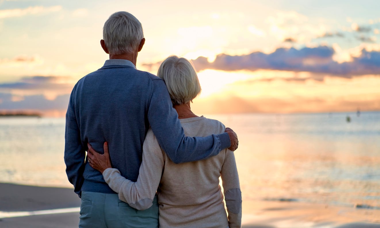 An elderly couple stands arm in arm on the beach, facing the ocean and watching the sunset—a quiet moment reflecting on the many Baby Boomer changes they’ve witnessed throughout their shared lifetime.