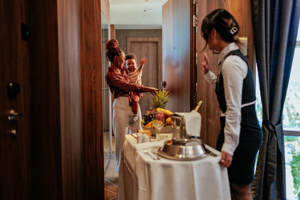 A hotel staff member delivers room service to a smiling woman holding a baby at the door, evoking the warmth and personal touch of things hotels used to offer guests. The baby waves excitedly while the staff waves back. A food tray with fruit and drinks is on a table in the foreground.