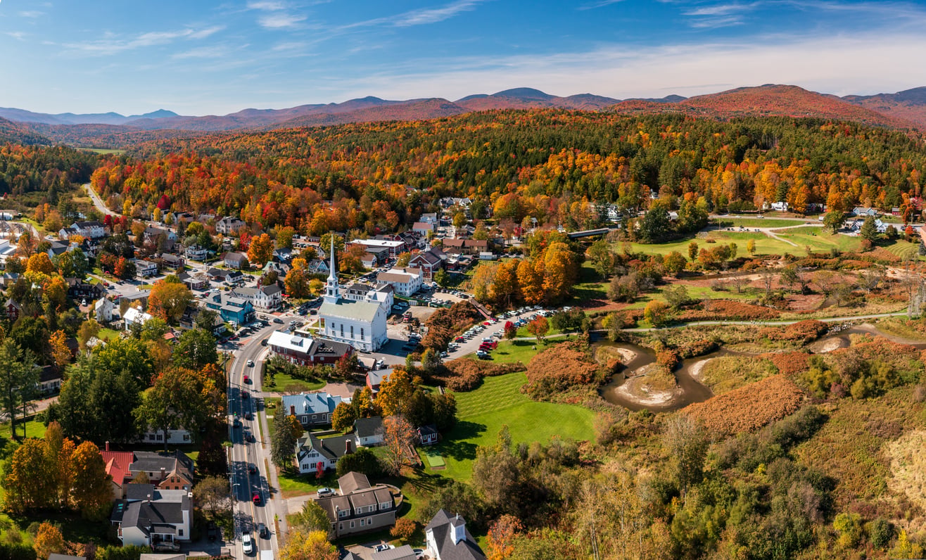 Aerial view of a small town surrounded by vibrant autumn trees, buildings clustered near a white church, with mountains and a clear blue sky in the background—an idyllic scene reminiscent of the best fall towns in America.