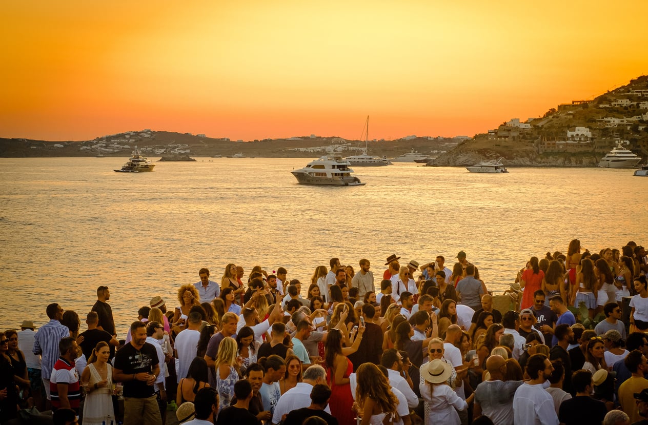 A crowd gathers by the sea at sunset, with yachts on the water and hills in the background. The sky glows orange, creating a festive, summer atmosphere.
