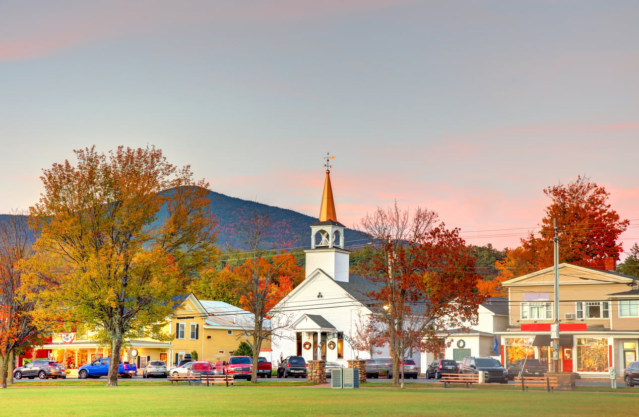 A small town in autumn—with colorful trees, a white church with a steeple, and nearby shops and houses—captures the charm found in the best fall towns in America; cars are parked along the street, and mountains rise in the background.