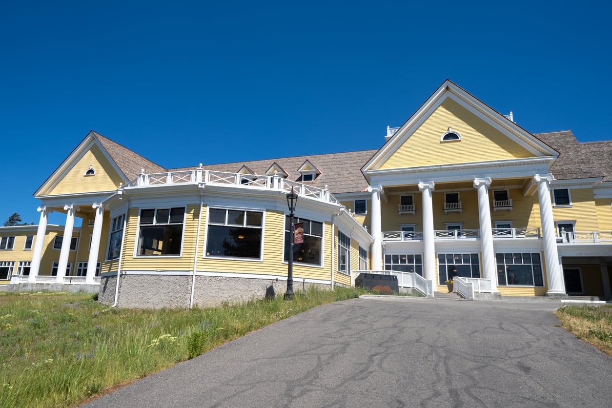 A large yellow building with white trim, columns, and a wraparound porch sits under a clear blue sky—a picture-perfect example of the elegance found in many Historic Hotels. The asphalt driveway in front leads up to the grand entrance, while manicured grass borders the building and driveway.