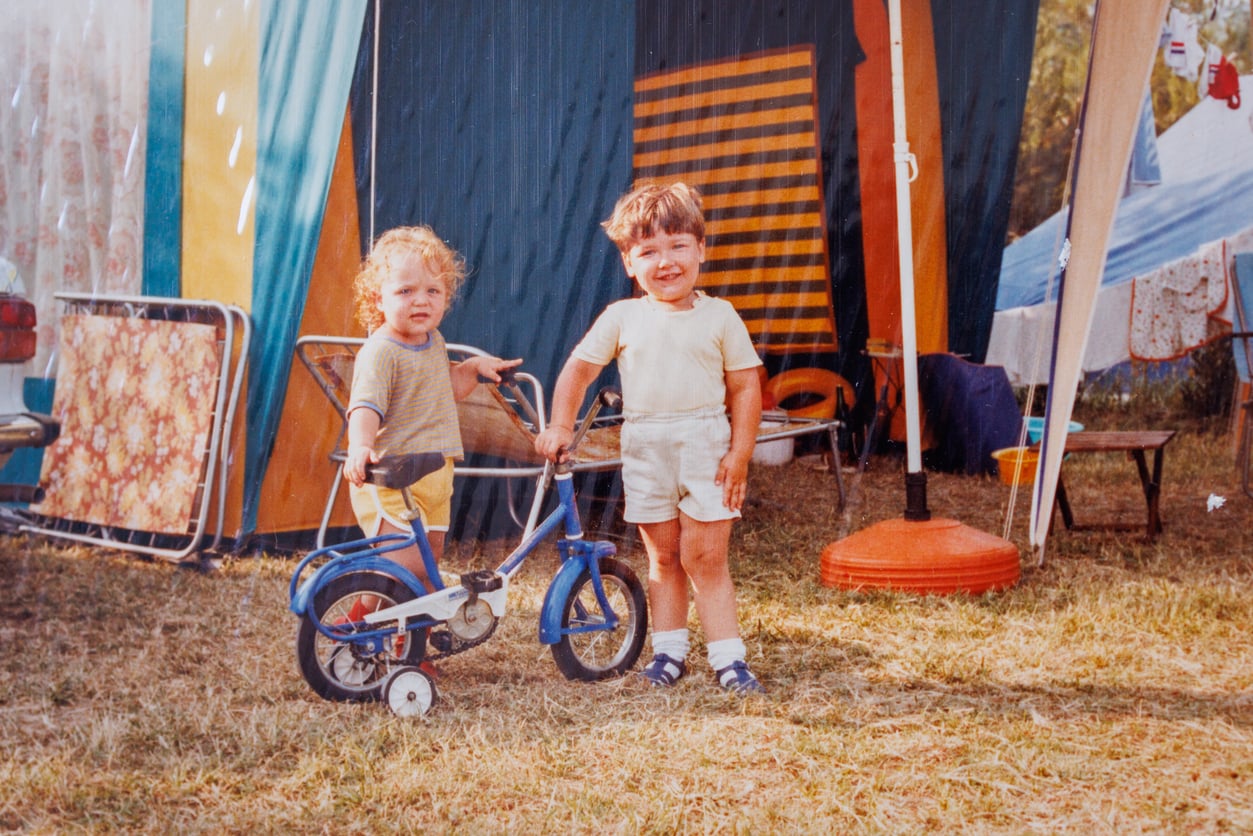 Two young children stand outside on grass by a blue tricycle. They are in front of colorful tents and camping equipment. One child is smiling; the other holds the tricycle. It appears to be a family camping trip.