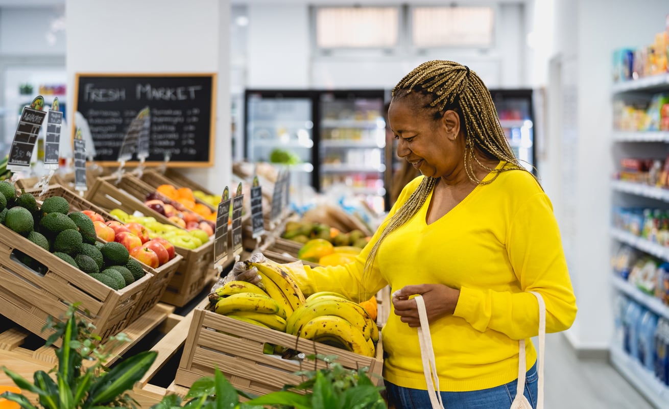 A woman in a yellow sweater smiles while selecting bananas at a grocery store. She holds a reusable bag and stands near wooden crates filled with fruits and vegetables.