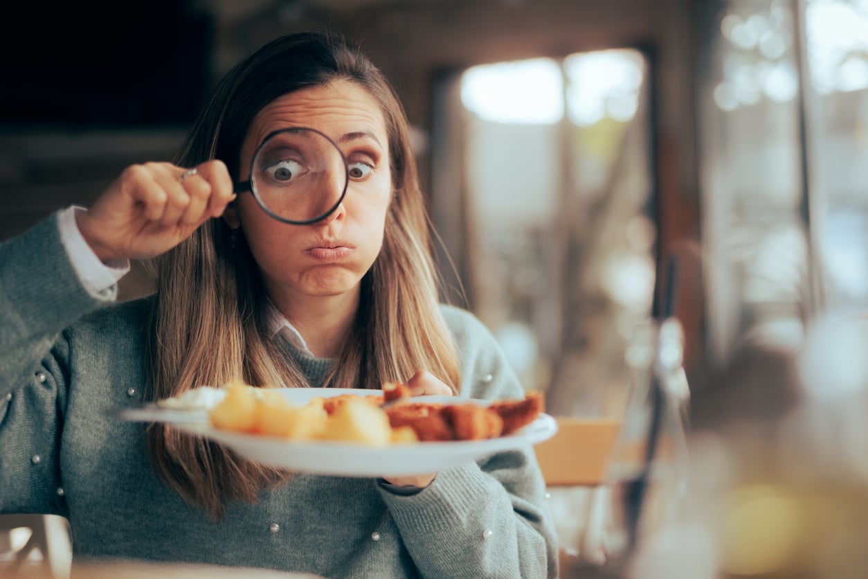 A woman holding a magnifying glass to her eye looks at a plate of food with a funny expression, puffing her cheeks, in a bright indoor setting.