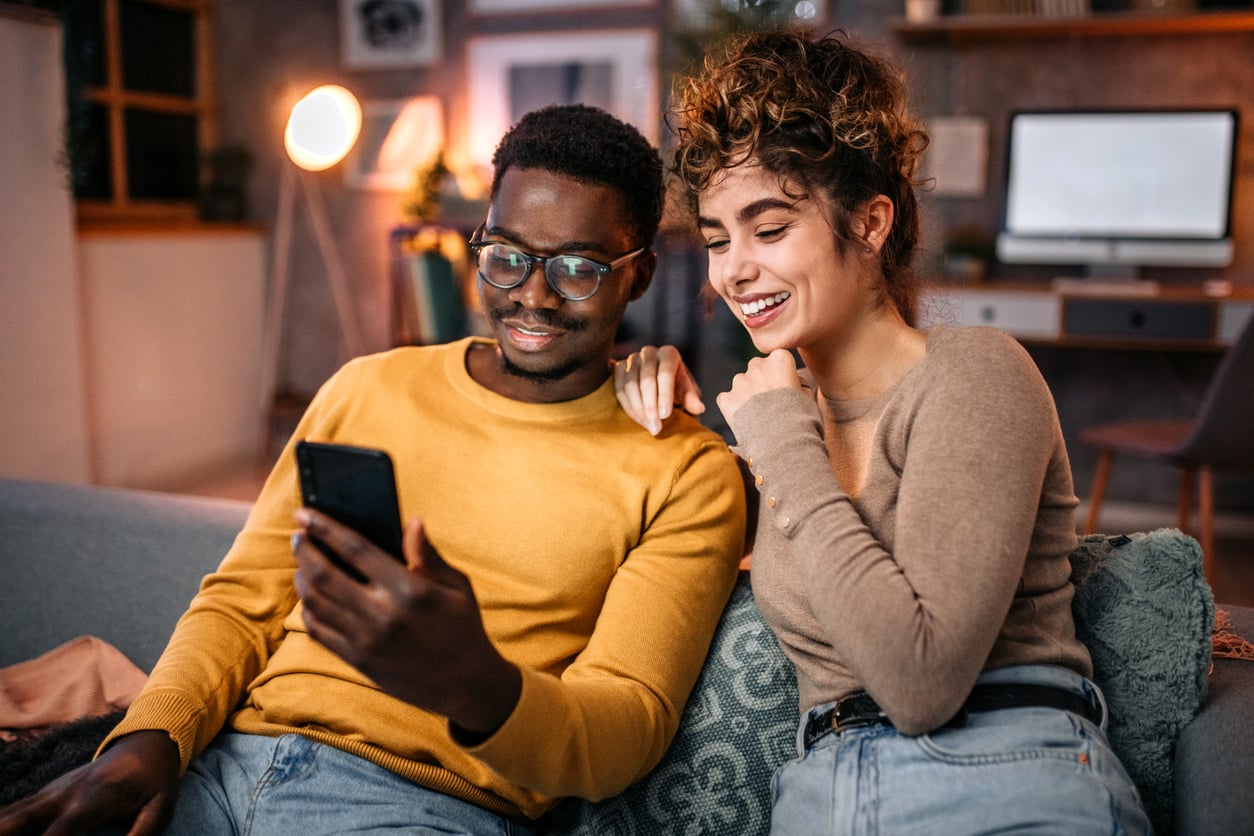 A man and woman, reflecting on Baby Boomer changes, sit together on a couch, smiling and looking at a smartphone in a cozy, softly lit living room.