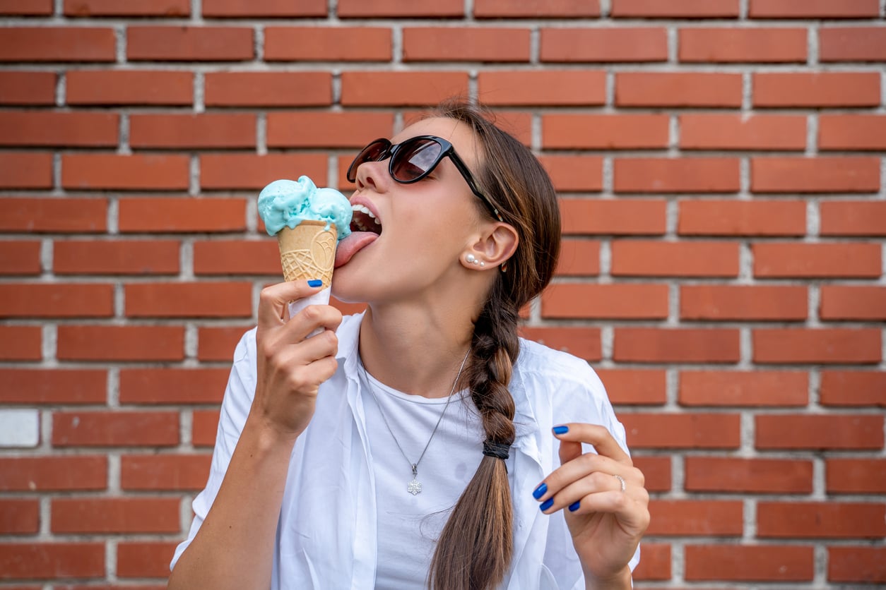 Woman with a braid and sunglasses prepares to lick a blue ice cream cone, standing in front of a brick wall. She wears a white shirt and has blue painted nails.
